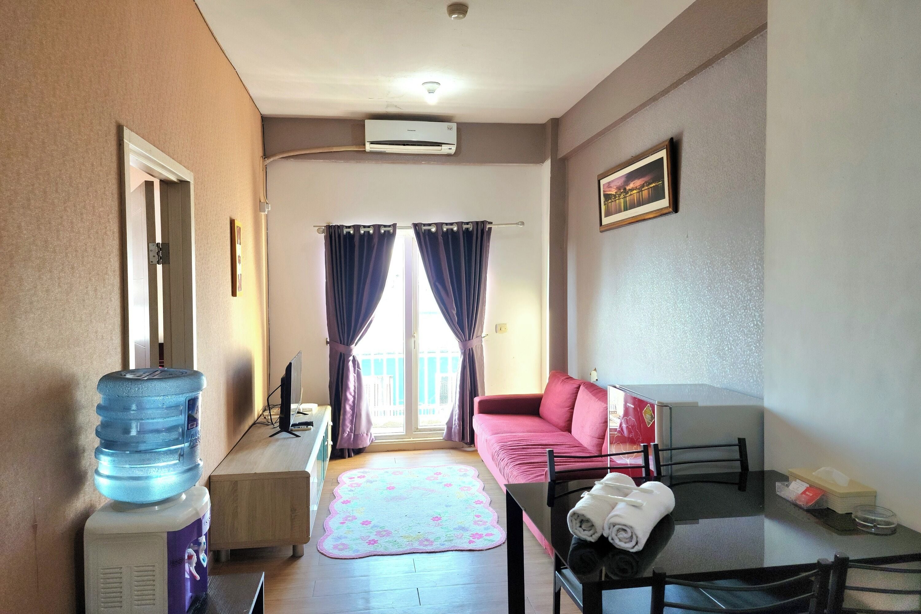 A compact living space with a sofa, a table with a chair, a water dispenser, and a window with curtains.