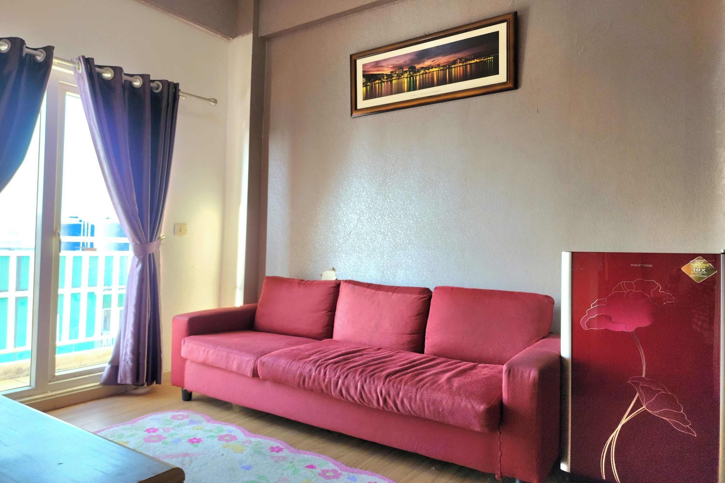 A red sofa in a room with a large window, a framed picture on the wall, and a refrigerator with a flower design.