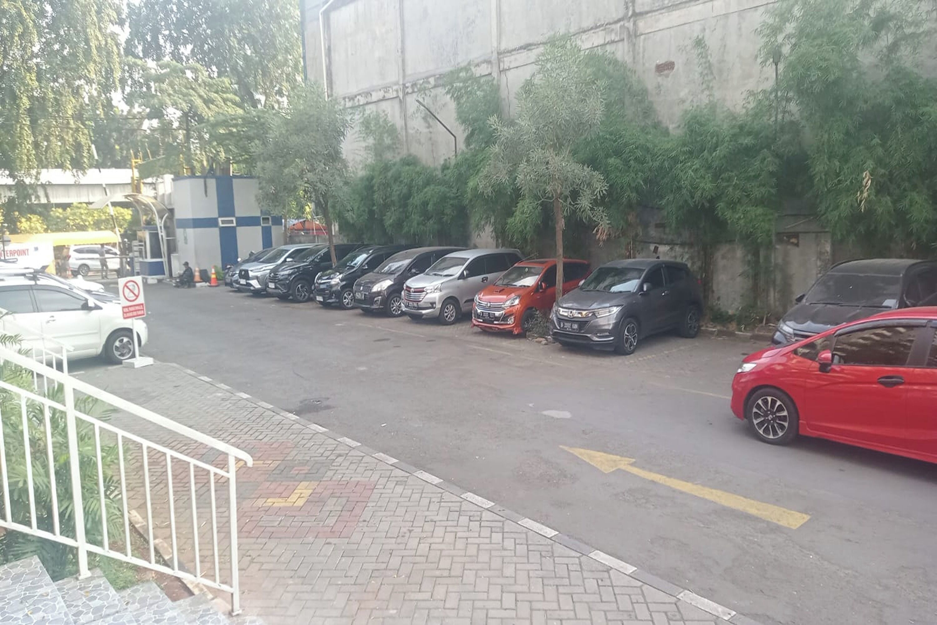 Um estacionamento com vários carros estacionados em fileiras, uma escadaria que leva a um andar superior e um edifício com fachada de concreto ao fundo.