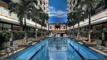 A swimming pool with a central slide, surrounded by palm trees and a multi-story building with balconies.