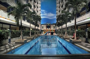 A swimming pool with a central slide, surrounded by palm trees and a multi-story building with balconies. - Queen Room 2 @ Grand Center Point Apartment (Bekasi)