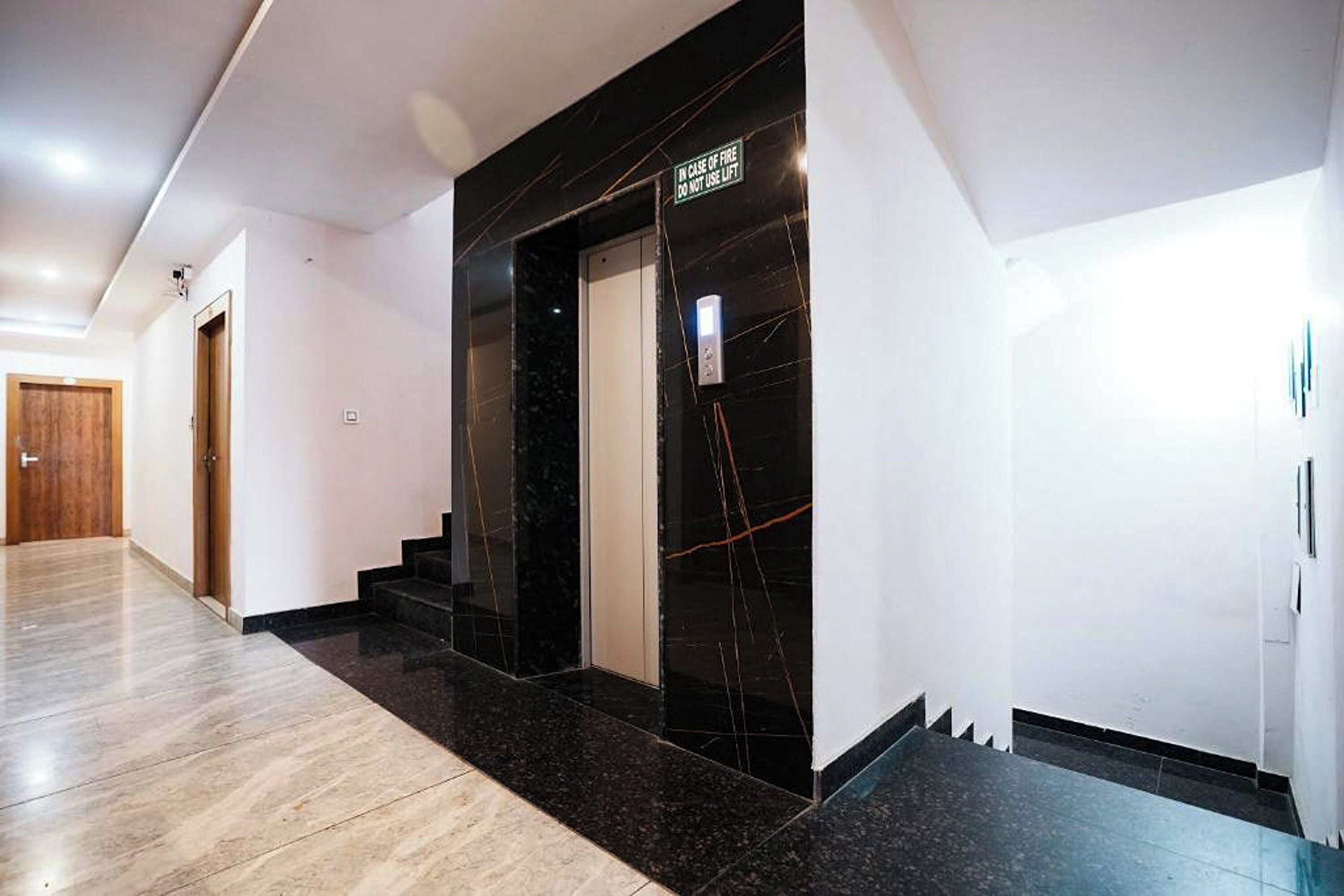 A modern hallway with a marble floor, a black elevator, and a wooden door at the end.