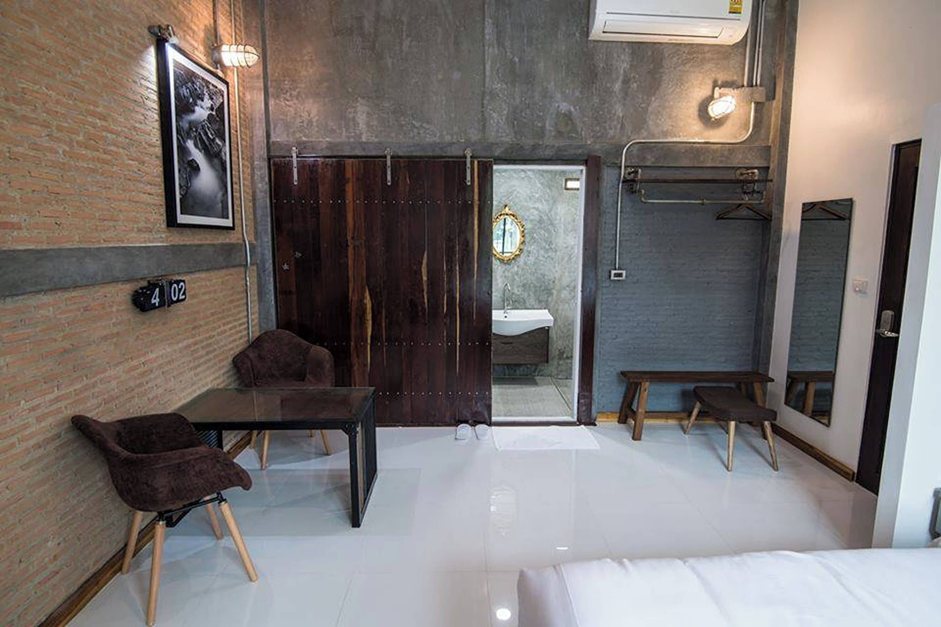 A modern hotel room with a brick wall, a wooden door, a glass shower enclosure, a white sink, a bed, a small table, and two chairs.