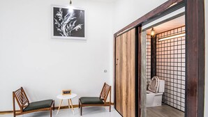 Une pièce comprenant deux chaises en bois, une petite table ronde, un tableau encadré accroché au mur et une porte coulissante donnant sur une salle de bain avec des toilettes et un lavabo.