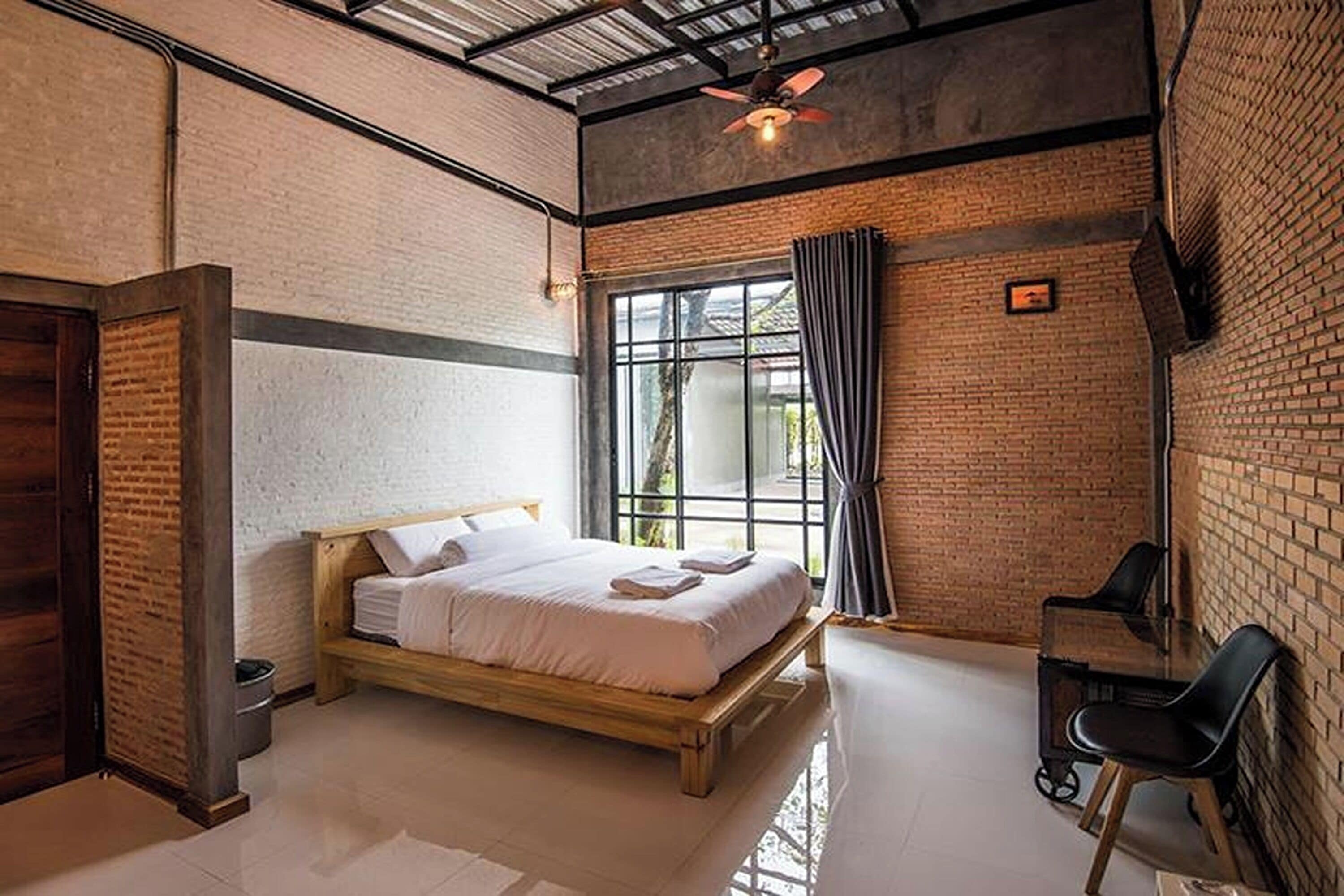 A bedroom with a wooden bed, a desk, and a chair, featuring exposed brick walls and a large window with curtains.