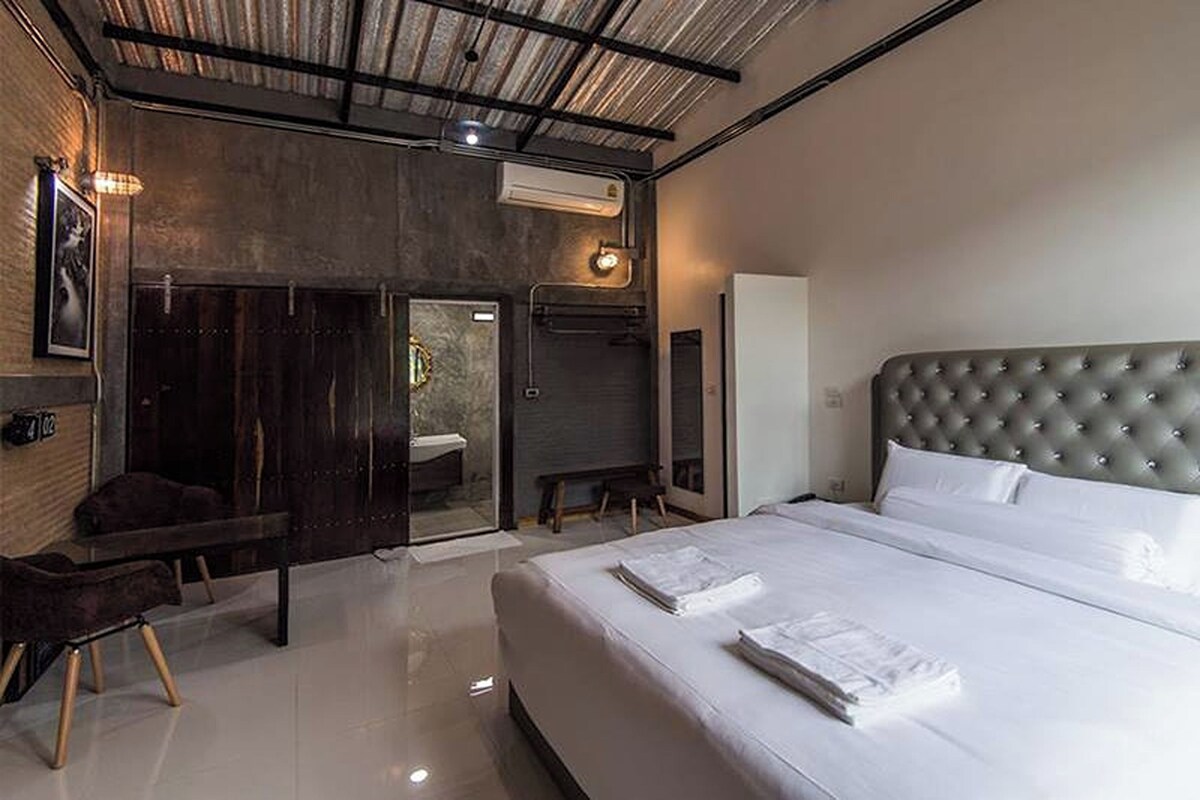 Une chambre d’hôtel moderne dotée d’un grand lit, d’une porte en bois foncé, d’un petit coin salon et d’une salle de bain visible à travers une paroi en verre.