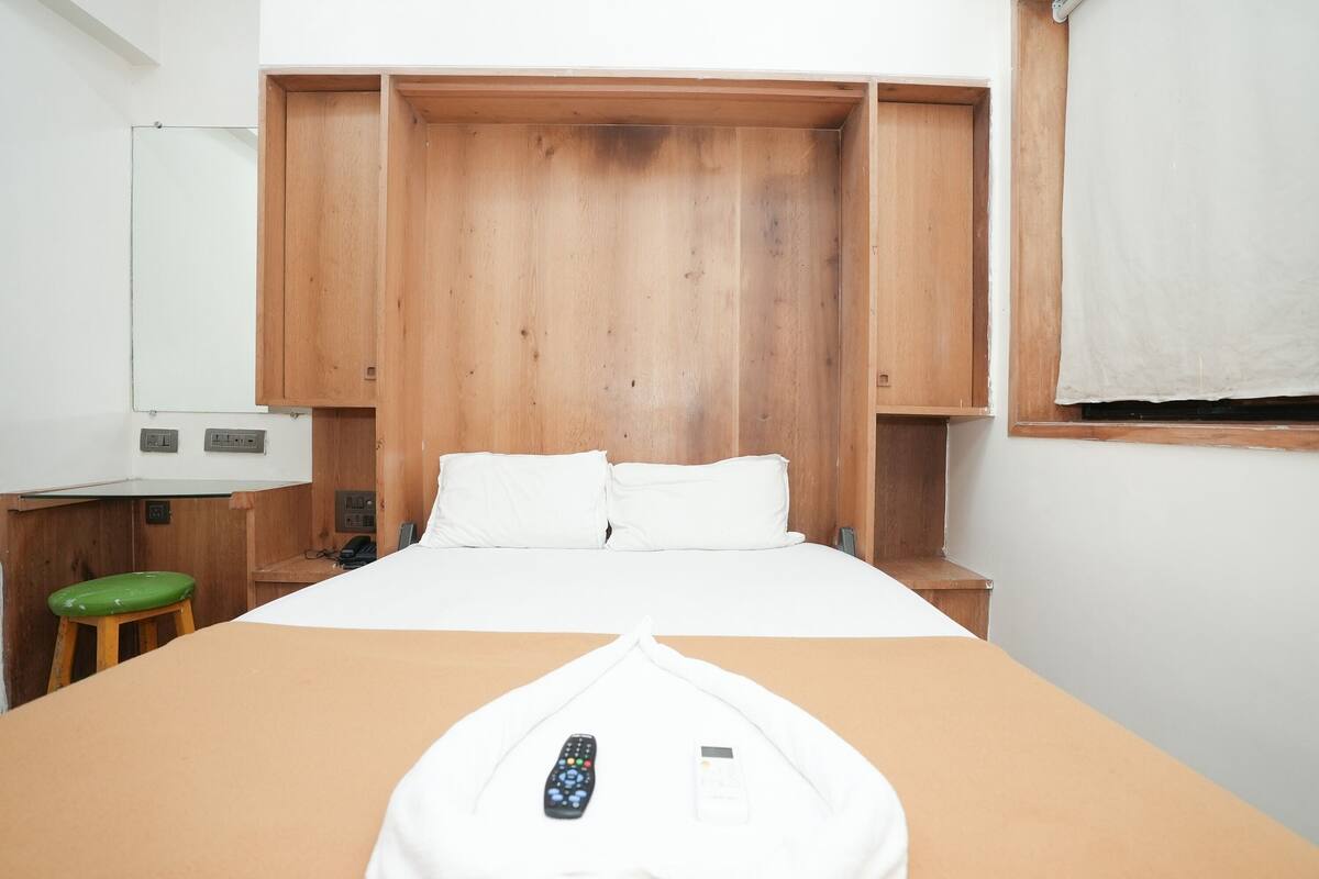 A hotel room with a bed, wooden wardrobe, a green stool, a mirror, and a window with a white curtain.