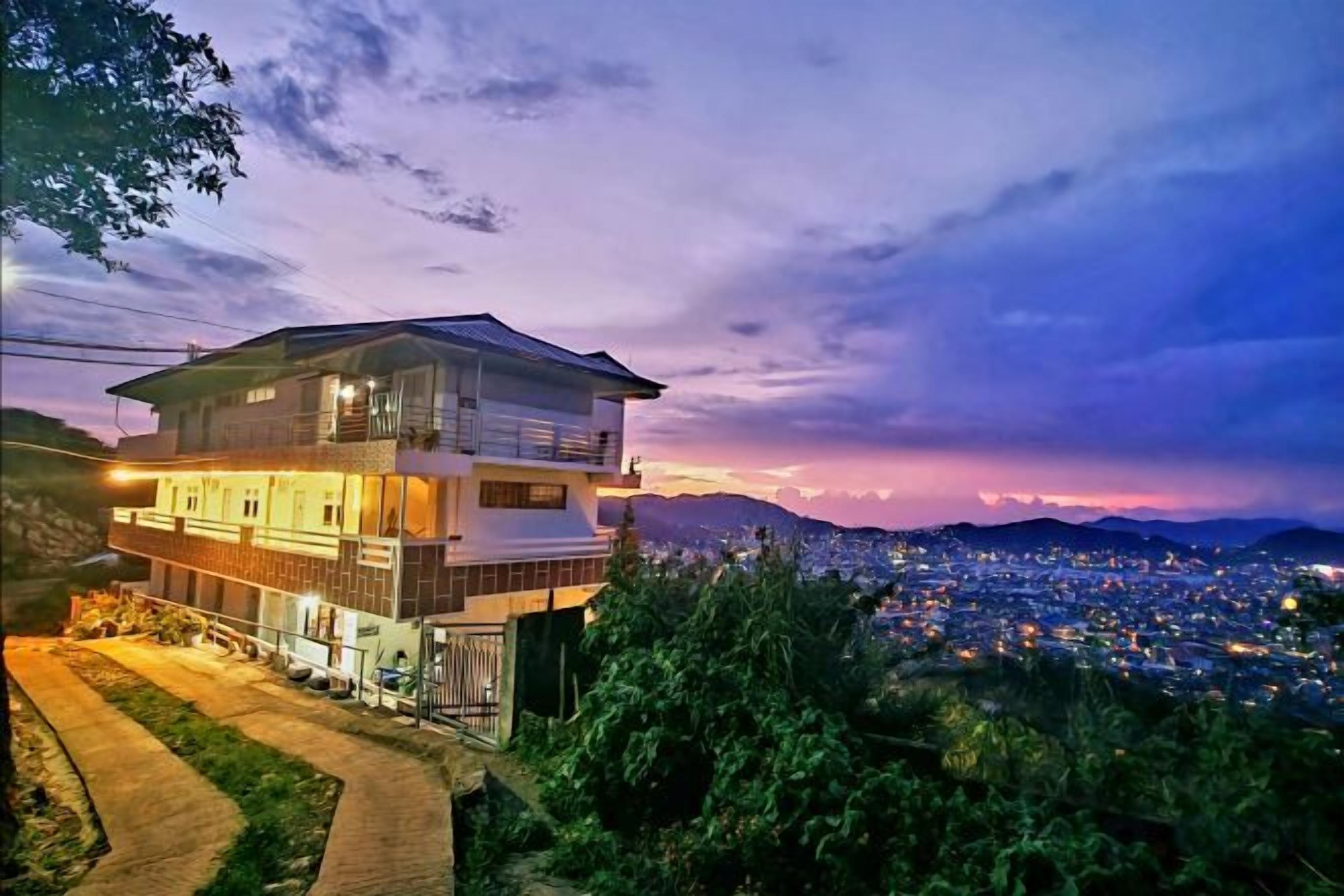 A multi-story building with a balcony, surrounded by greenery and overlooking a cityscape at dusk.