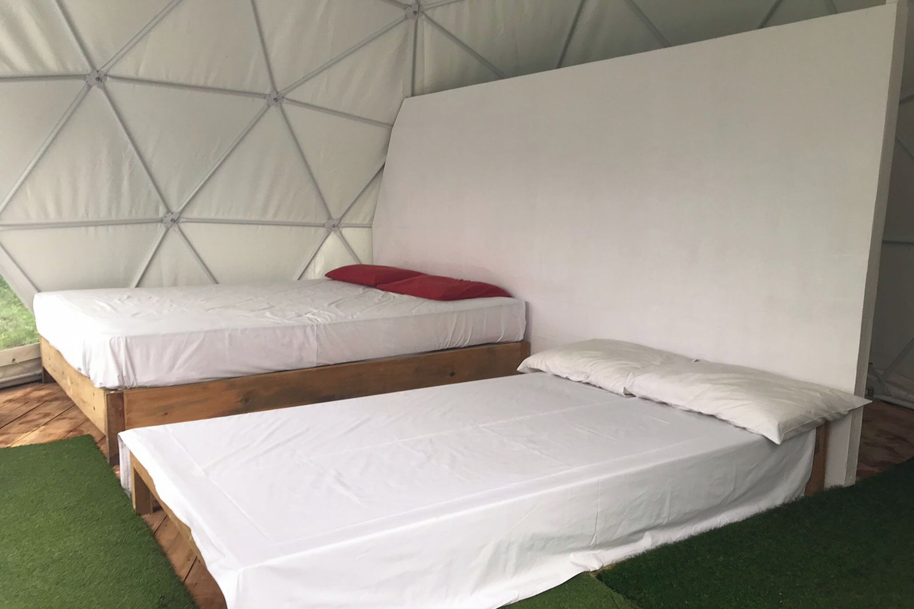 Deux lits simples avec une literie blanche et un oreiller rouge, des têtes de lit en bois et, au fond, une structure évoquant une tente.