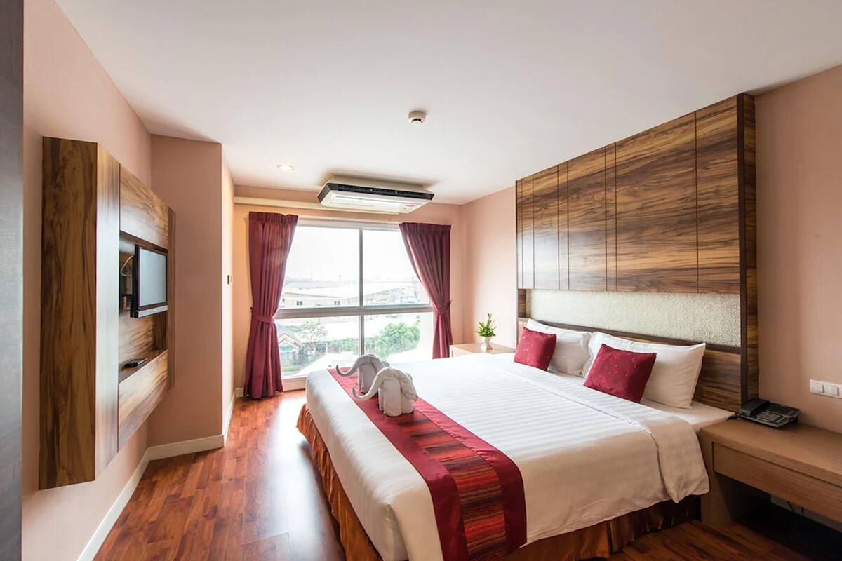 A hotel room with a large bed, a TV mounted on the wall, a wooden headboard, and a window with curtains.