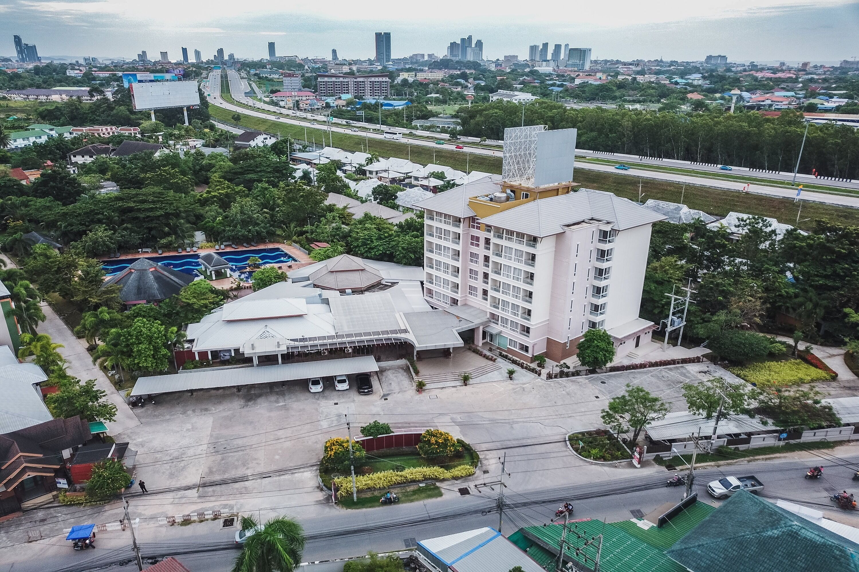 An aerial view of a hotel with a large parking lot, a swimming pool, and a landscaped garden area.