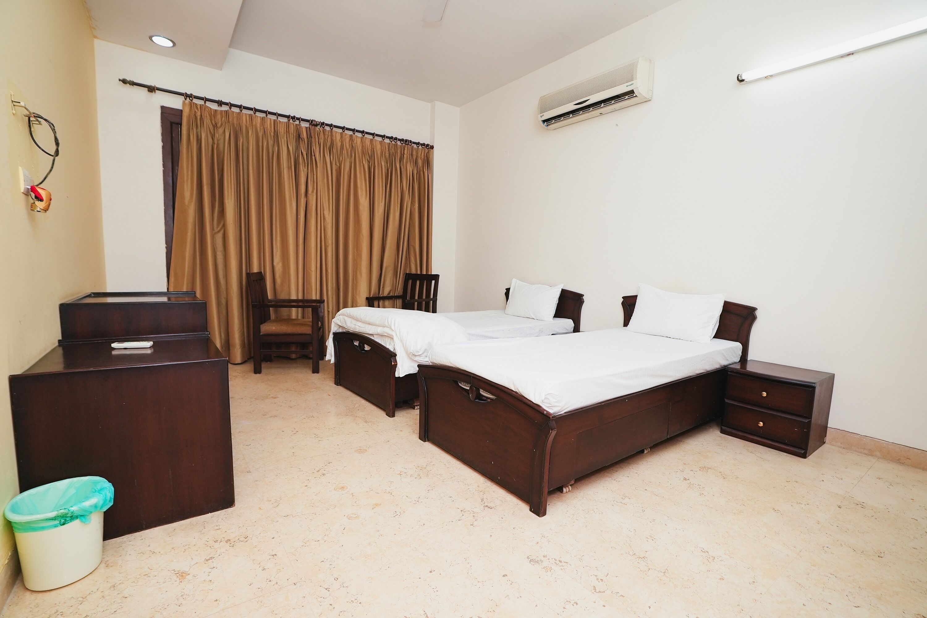 A bedroom with two beds, a desk, a chair, a trash bin, and an air conditioning unit.