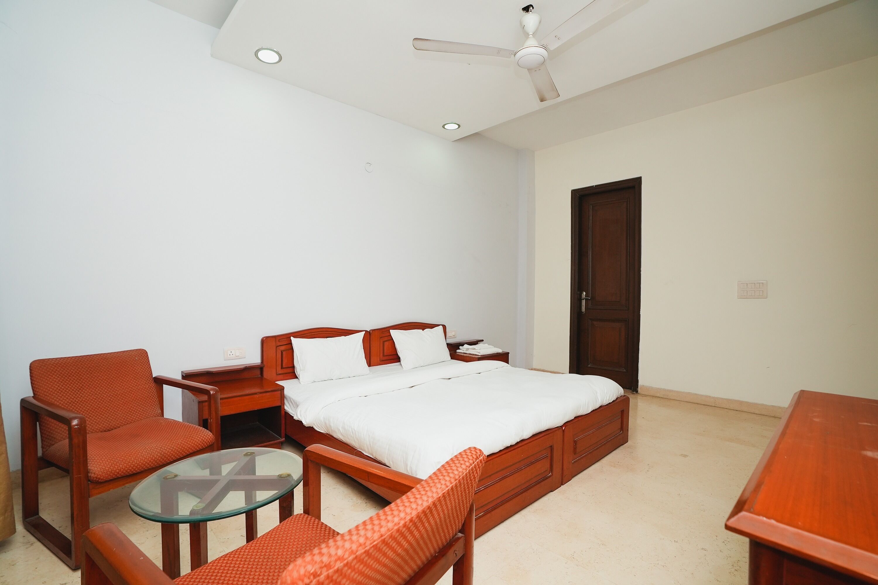 A bedroom with a bed, two wooden chairs, a glass table, a ceiling fan, and a wooden door.