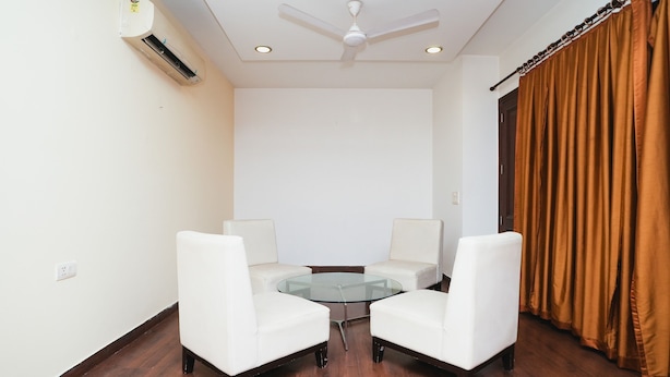 A room with a ceiling fan, a glass table surrounded by two white chairs, and a brown curtain on the right. - Home 92 (Noida)