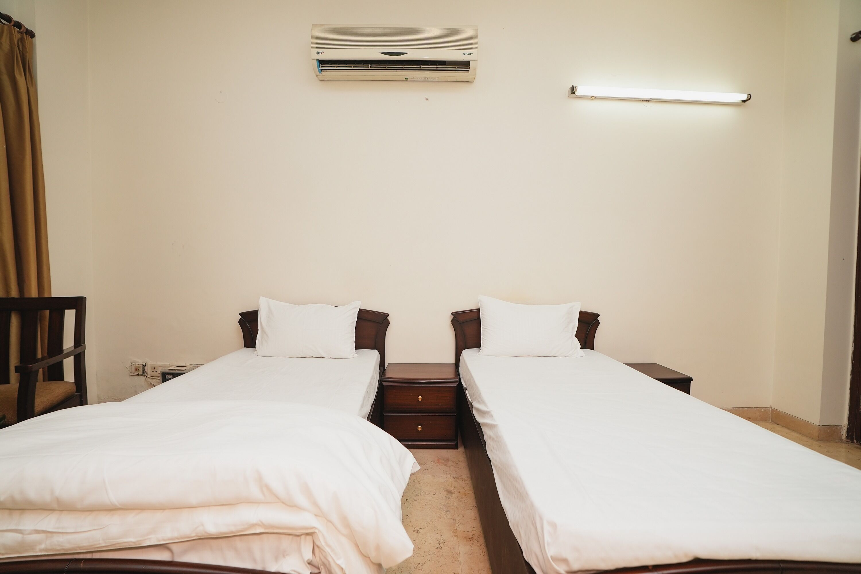 A room with two single beds, a bedside table, a wall-mounted air conditioner, and a bedside lamp.