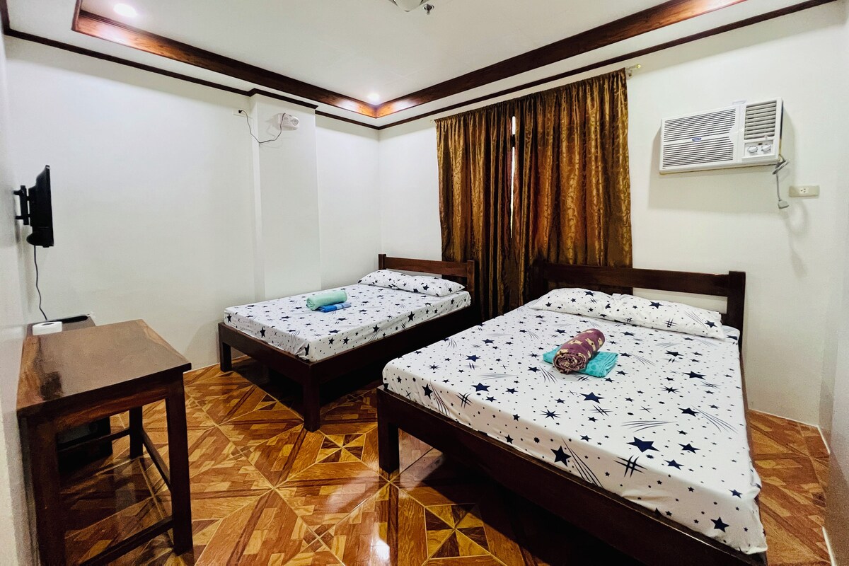 A room with two beds, a desk, a ceiling light, an air conditioner, and a TV mounted on the wall.