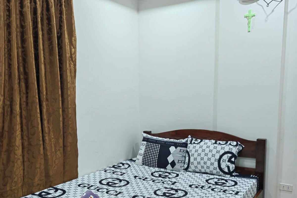 Una piccola camera con un letto singolo, una coperta a motivi, un poggiatesta in legno e una tenda a sinistra.
