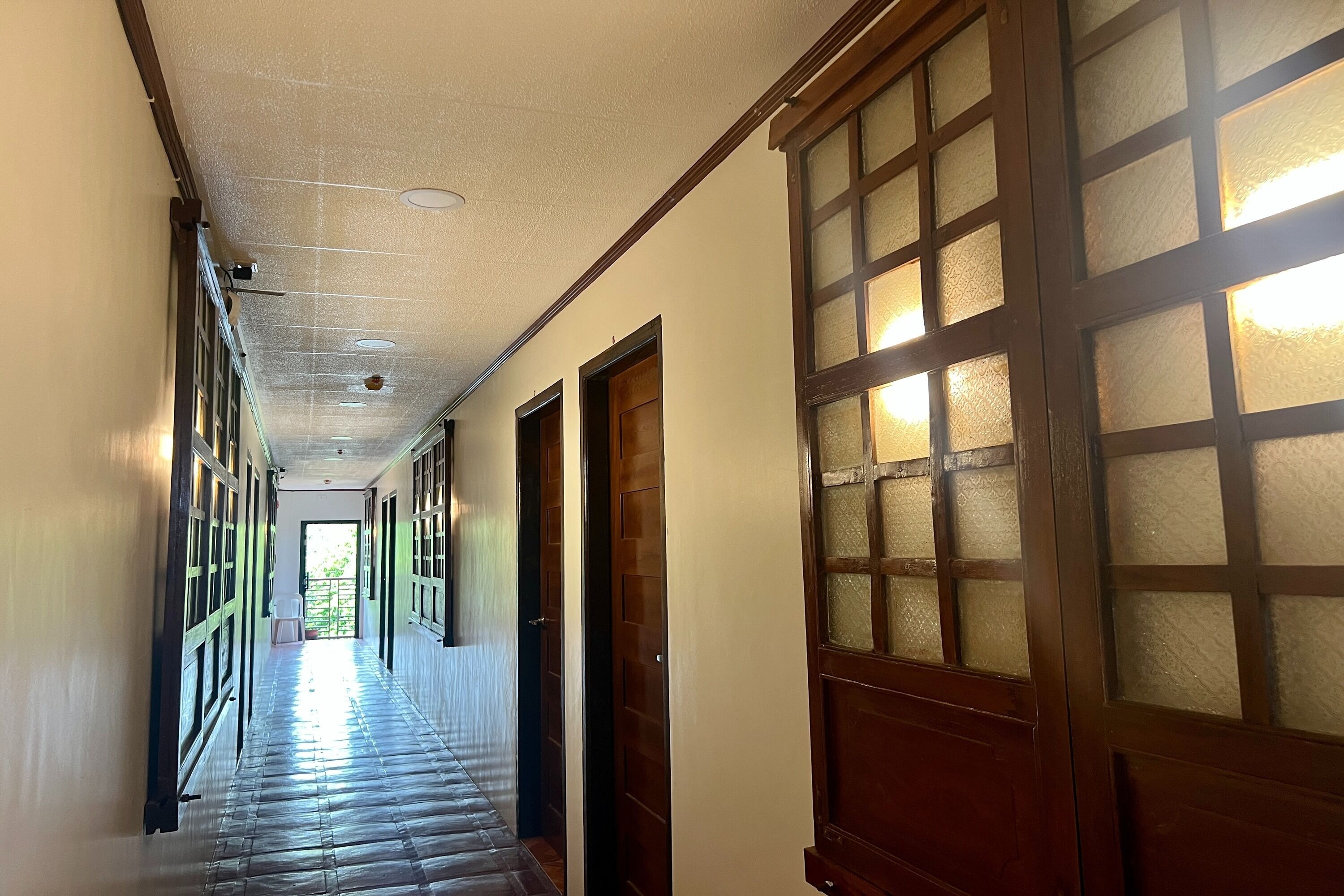 A long corridor with wooden doors and windows, leading to a bright area.