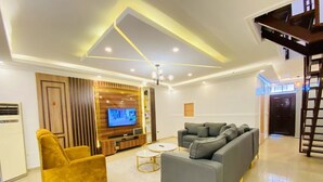 A modern living room with a grey sofa, a mustard-colored armchair, and a TV. - Deluxe Apartment (Lagos)