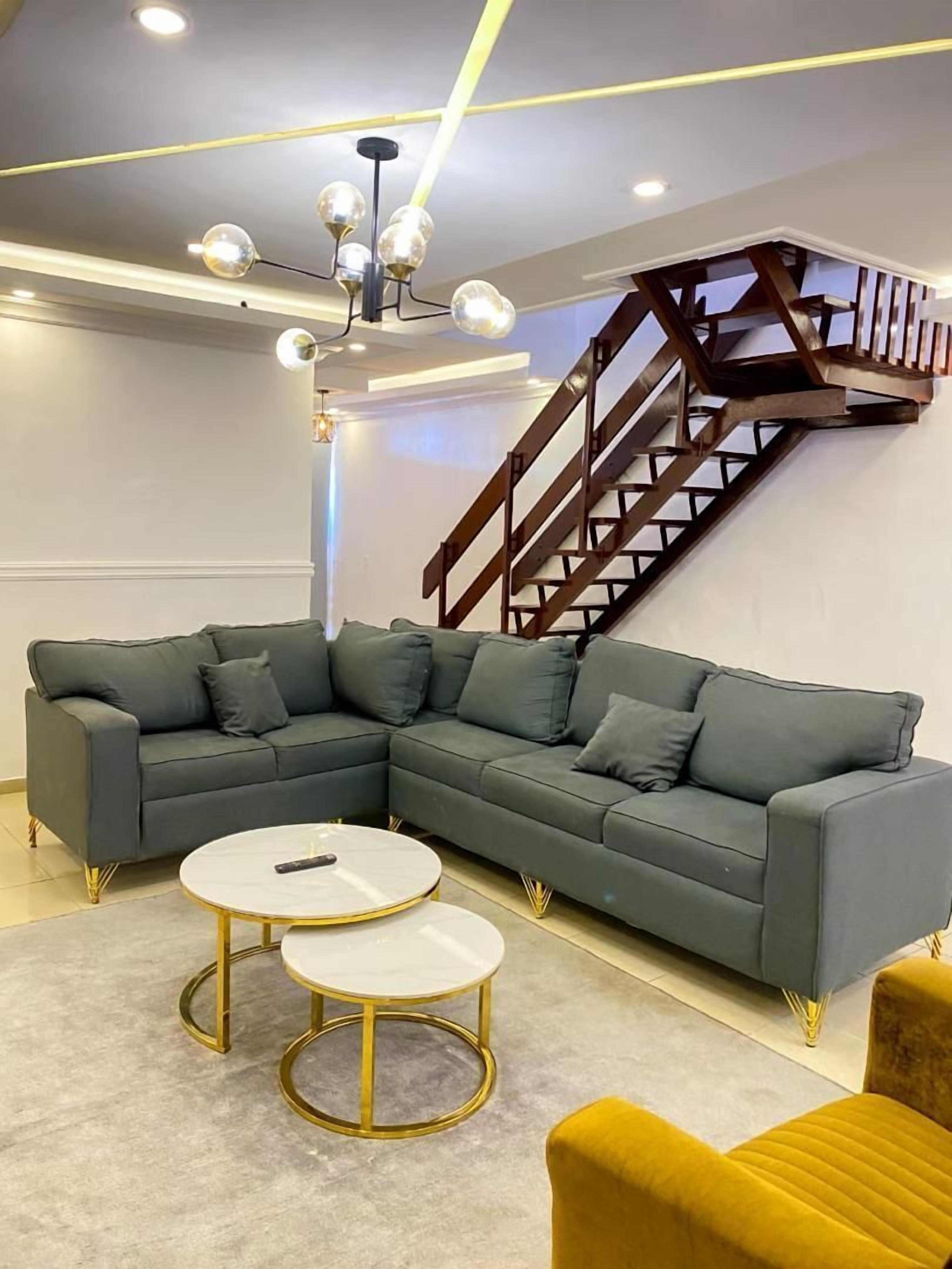 Un salon moderne avec un canapé d’angle, deux petites tables rondes, un fauteuil jaune et un escalier en bois.