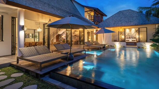 A swimming pool with lounge chairs and umbrellas, a staircase leading to a balcony, and a well-lit interior. - Villa Nusantara 5 by Alfred in Bali (Canggu)