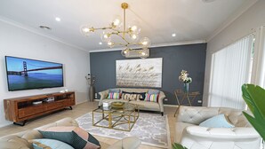 A modern living room with a large flat-screen TV, a tufted sofa, a coffee table, and a chandelier. - Hallam 5BRM Rental Entire House (Hallam)