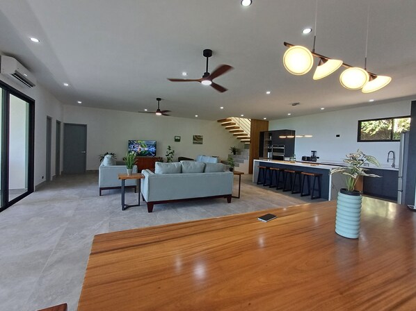 A modern living room with a sofa, a wooden coffee table, and a TV. There is a kitchen area with a bar and stools. - Hear the waves from either pools or patios, or walk to the beach, ocean views (Manuel Antonio)