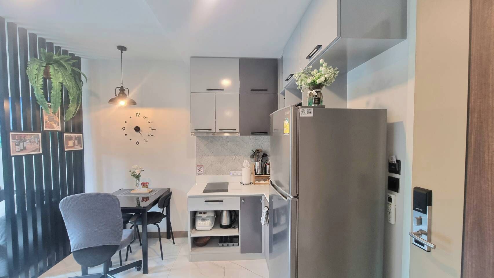 A compact kitchen with a refrigerator, a dining table with chairs, a hanging plant, and a small countertop with a coffee machine.
