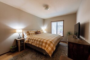 A bedroom with a bed, bedside lamps, a television on a wooden stand, a window with blinds, and a small plant.