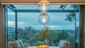 A dining area with a wooden table, two chairs, a vase with flowers, and a view of greenery through large glass doors. - Wren House Victor Harbor (Victor Harbor)