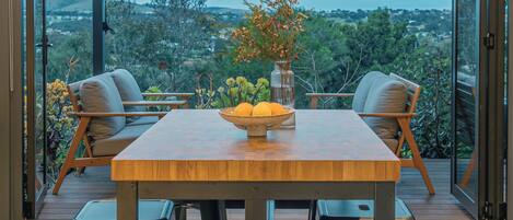 Un coin repas avec une table en bois, deux chaises, un vase de fleurs et une vue sur la verdure grâce à de grandes portes vitrées.