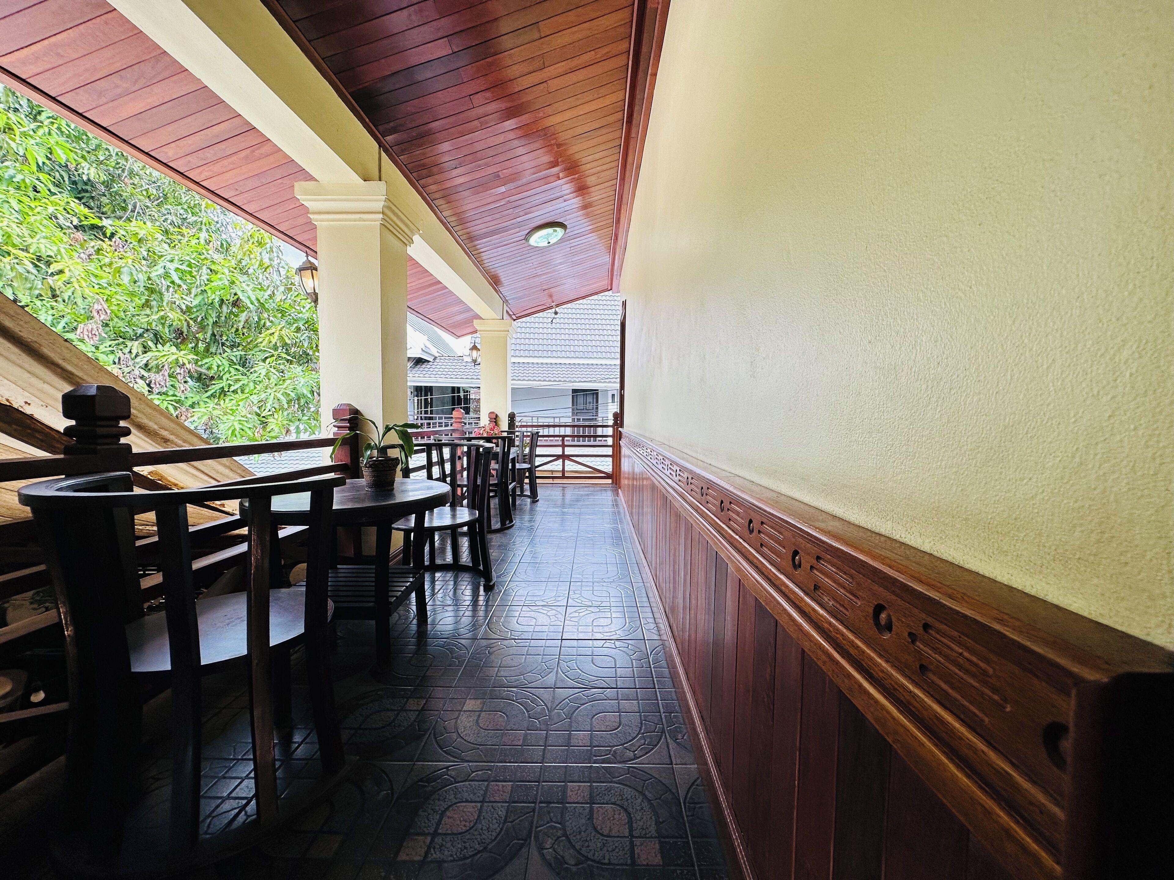 A covered outdoor seating area with wooden tables and chairs, a tiled floor, and a view of greenery and buildings in the background.
