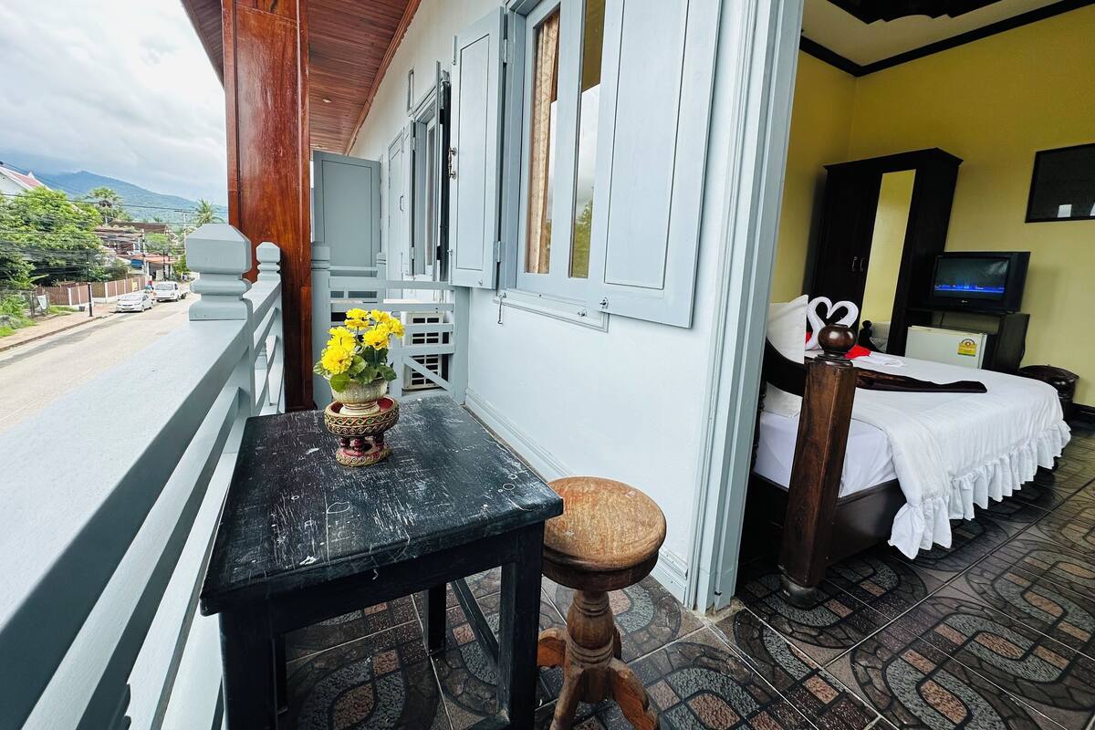 A balcony with a wooden railing, a small table with a vase of flowers, a bed with a white bedspread, and a television on the wall.
