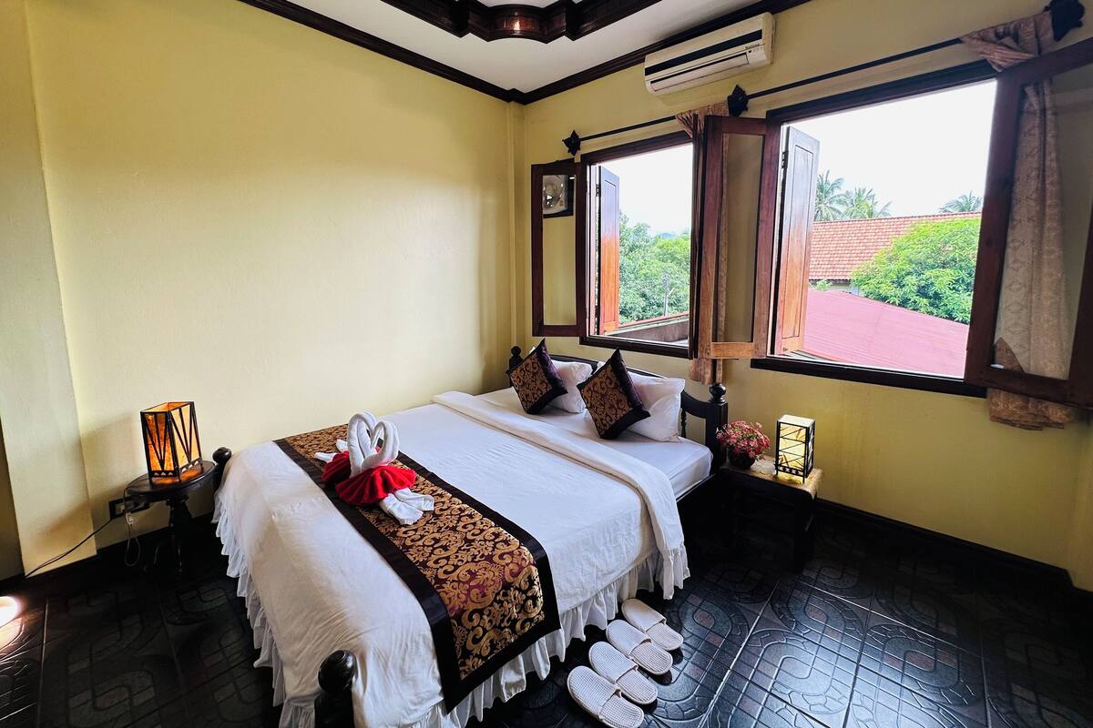 A hotel room with a bed, a window with an open view, a bedside table with a lamp, and slippers on the floor.