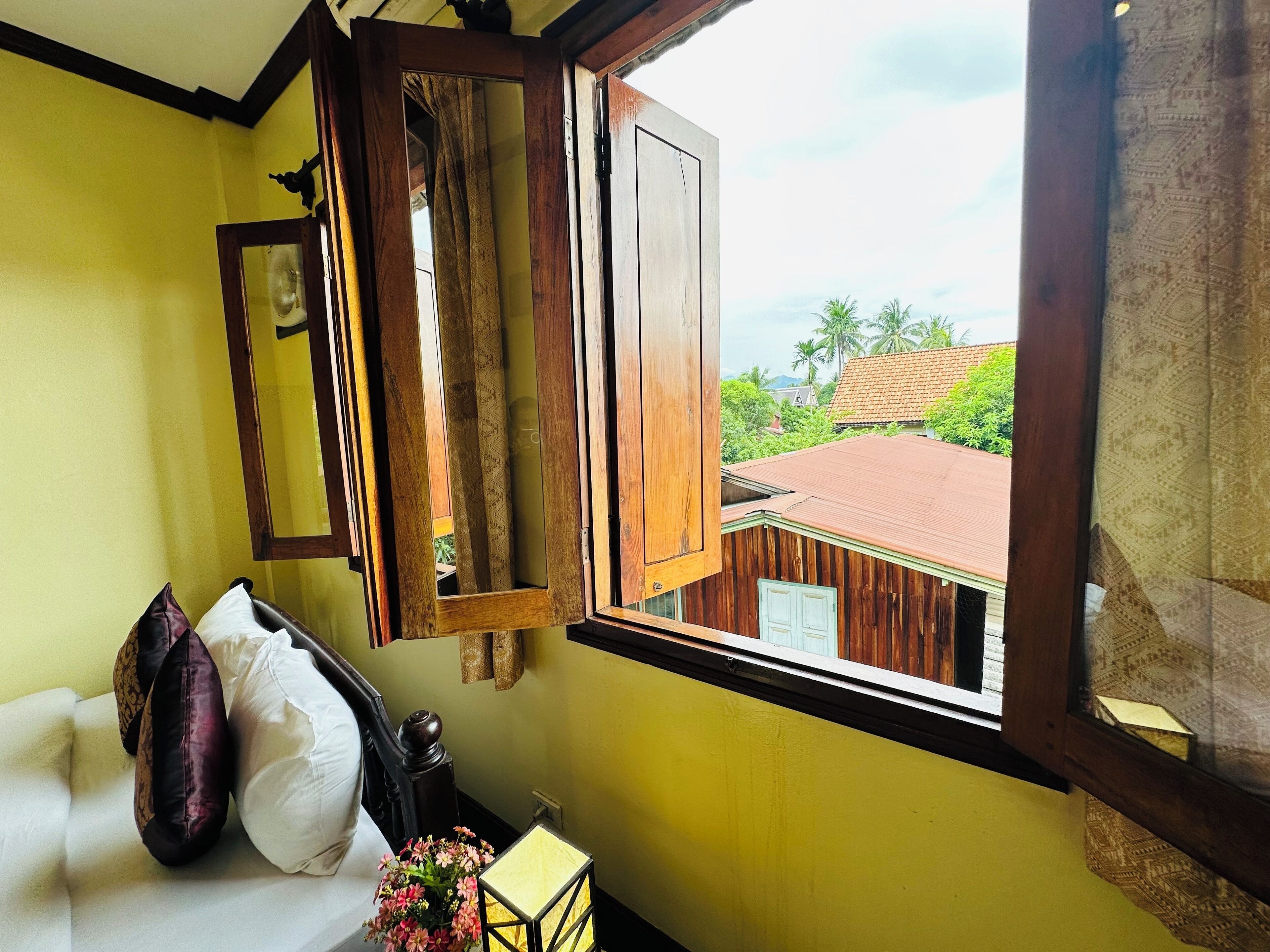 A room with a bed, a window with an open wooden shutter, a mirror, and a vase with flowers.