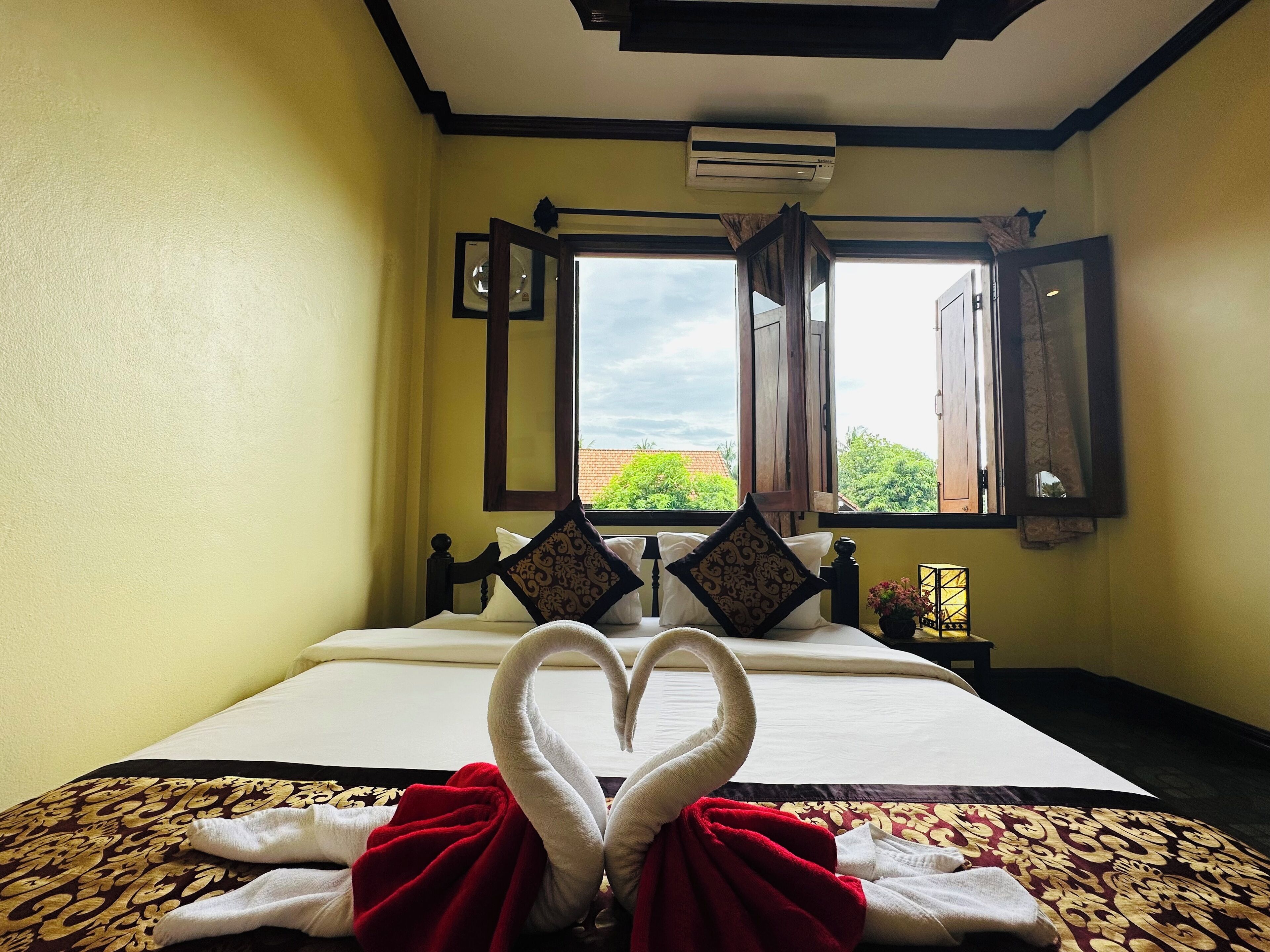 A hotel room with a bed, two windows, and a view of greenery outside.