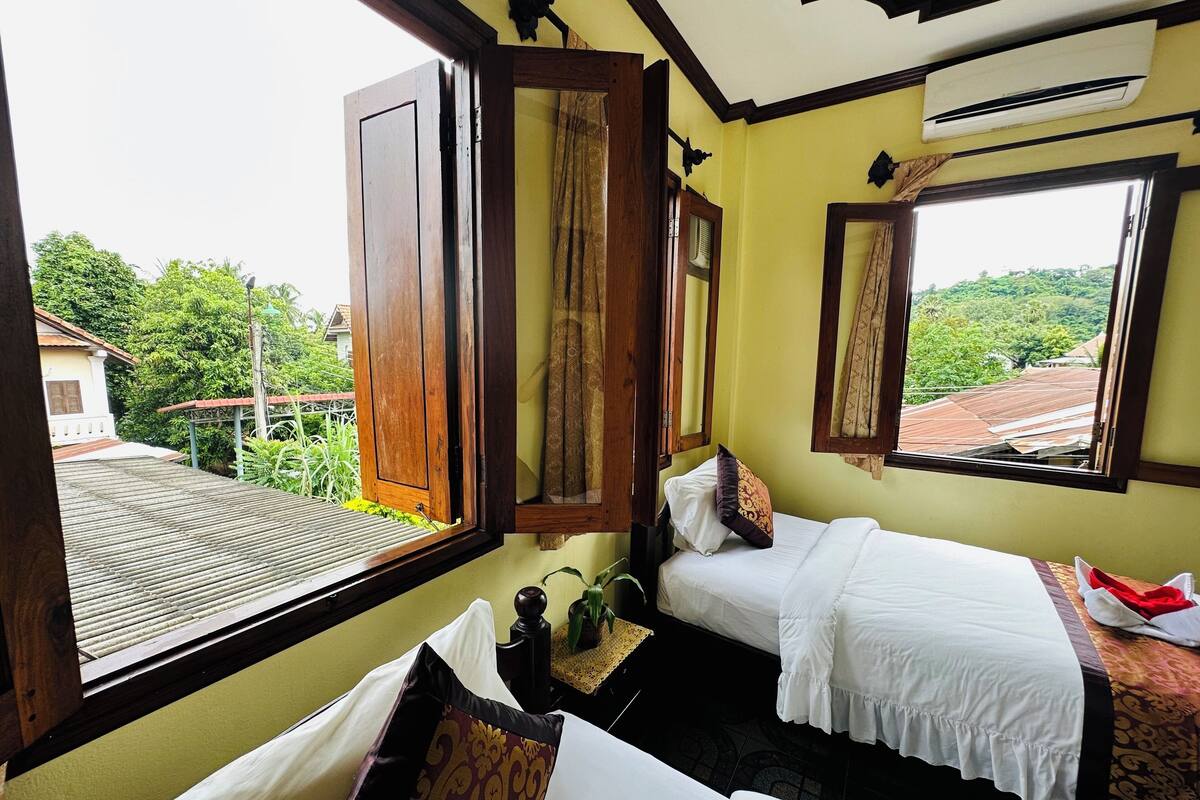 A room with two beds, a window with an open shutter, and a view of greenery outside.