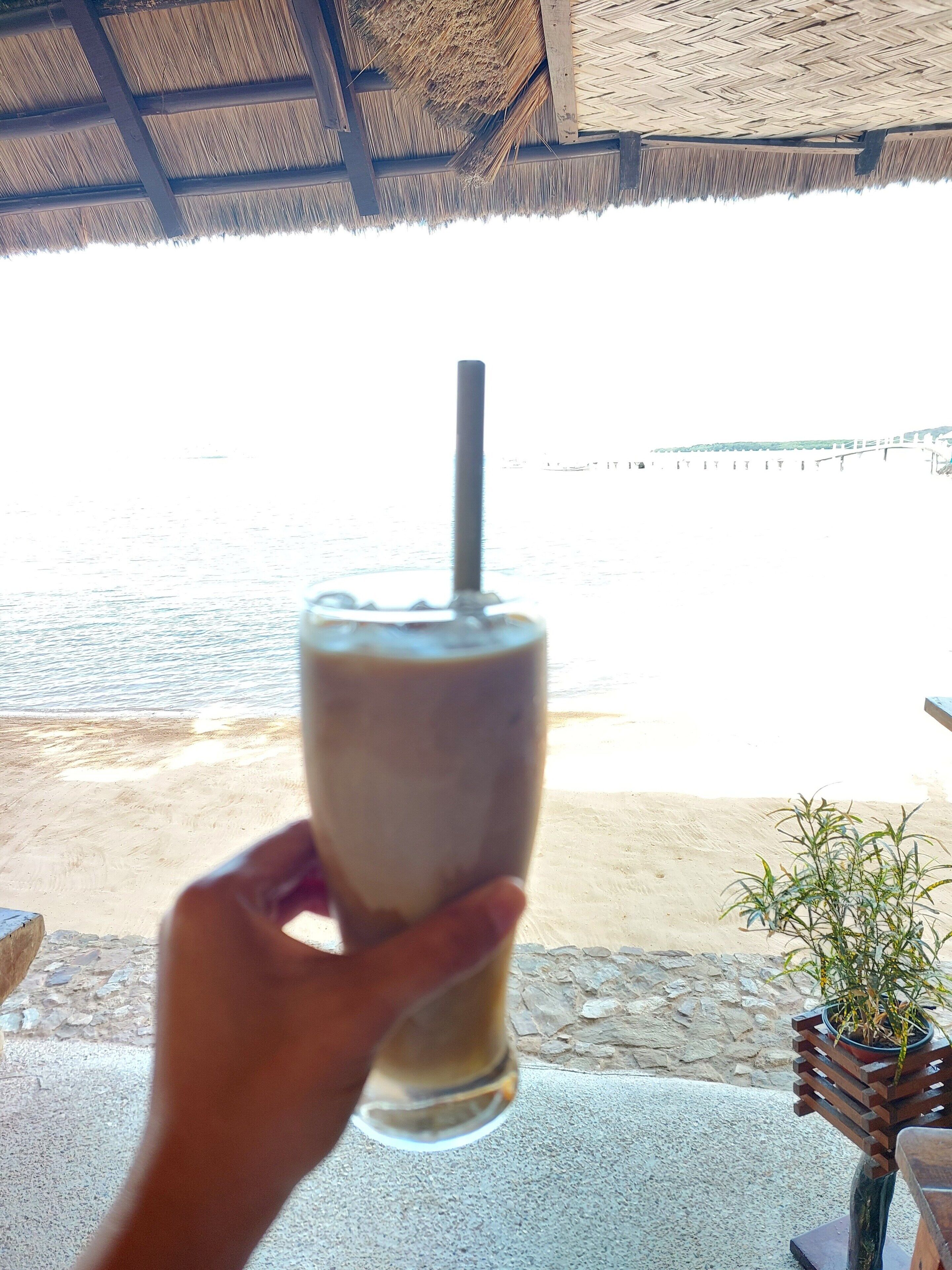 A hand holding a glass with a straw, a beach view, and a thatched roof structure.