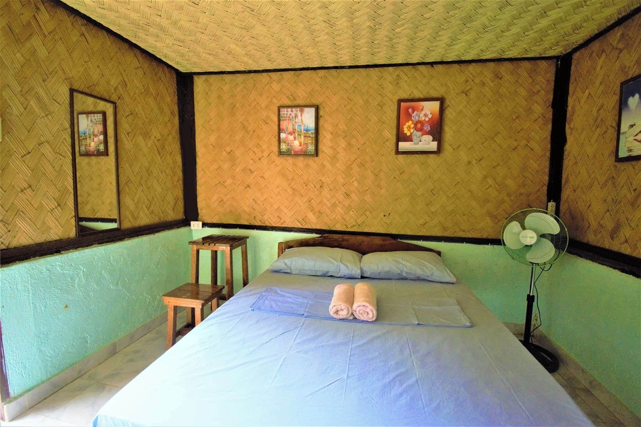 Une chambre avec un lit, deux oreillers, une table de chevet, un ventilateur sur pied et des tableaux encadrés accrochés au mur.