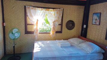 A room with a bed, a standing fan, a window with curtains, a wall-mounted picture, and a woven wall pattern.