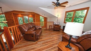 A spacious loft-style room with a wooden floor, leather sofas, a ceiling fan, and large windows offering a view of the outdoors. - Mountain Lodge • New Hot Tub • Great Mountain Views • Firepit • Pet Friendly (Marble)