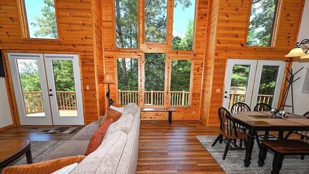 A cabin interior with a wooden floor, a sofa, a dining table with chairs, and large windows offering a view of the outdoors. - Mountain Lodge • New Hot Tub • Great Mountain Views • Firepit • Pet Friendly (Marble)