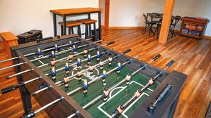 A foosball table in a room with wooden flooring, a table, chairs, and a cabinet. - Mountain Lodge • New Hot Tub • Great Mountain Views • Firepit • Pet Friendly (Marble)