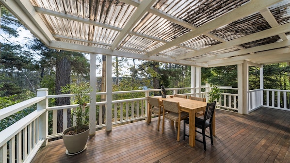 Una terraza techada al aire libre con mesa y sillas de madera, rodeada de barandillas blancas y plantas en macetas.