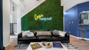 A modern living room with a black leather sofa, a neon sign reading "Come hang out!", and a green wall.