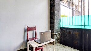 Ein Laptop steht auf einem kleinen Tisch neben einem hölzernen Stuhl in einer Ecke mit Fliesenboden und einem Metallgitter im Hintergrund.