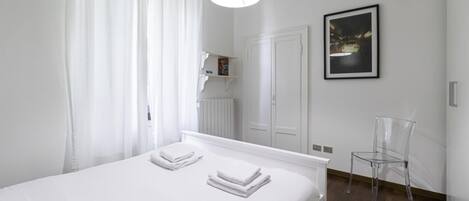 A clean, modern bedroom with a large bed, a bedside table, a chair, a mirror, and a framed picture on the wall.