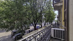 En balkon med gelænder af metal, udsigt over en gade med parkerede biler og træer.