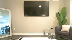 A modern living room with a flat-screen TV mounted on the wall, a white coffee table with a glass top, a gray sofa with a white pillow, and a large green plant. - SUAVE&SPACIOUS SUITE NEAR EVERYTHING (Atlanta)