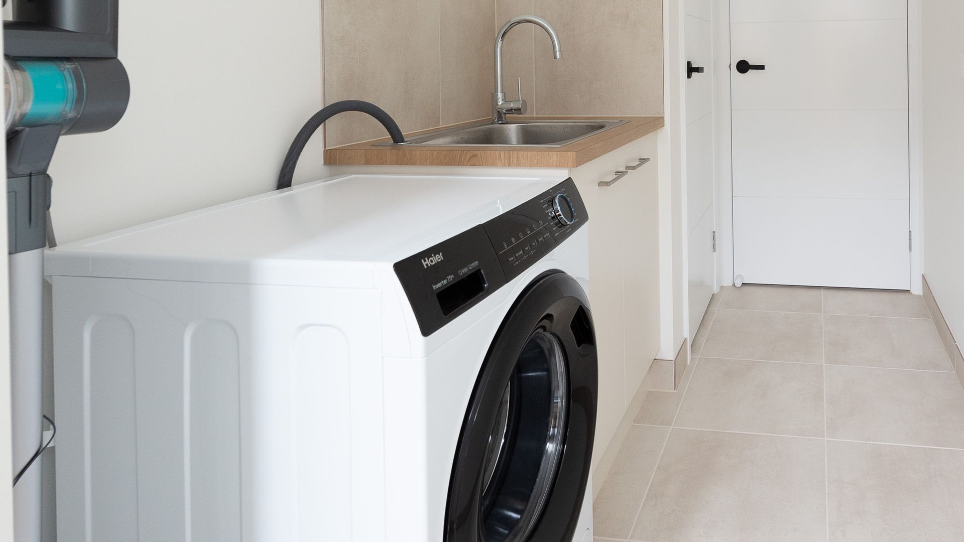 A compact, modern laundry room with a front-loading washer and dryer, a wooden countertop, a sink, and a door.
